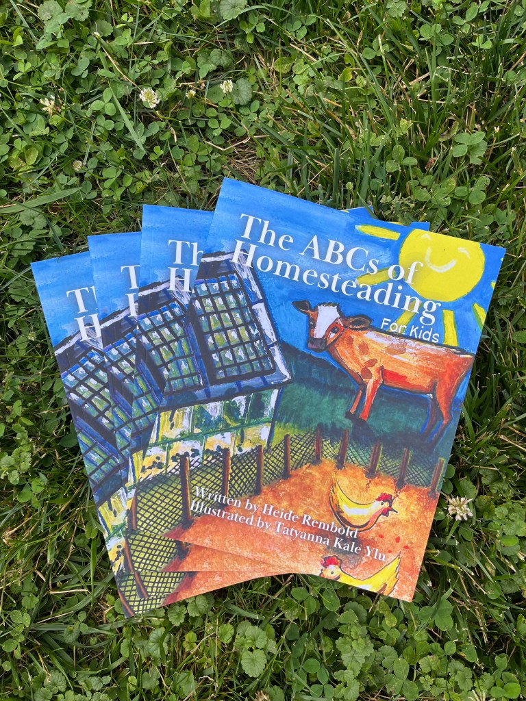 multiples copies of The ABCs of Homesteading for Kids stacked up on grass.